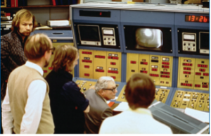 A group of men seated around a large console filled with buttons, monitors, and read-outs. On a small television screen, a circular blotch of white appears against a black background.