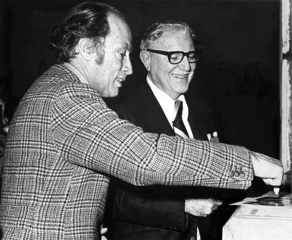 Two men point at a photo laying on a table, smiles clearly visible.