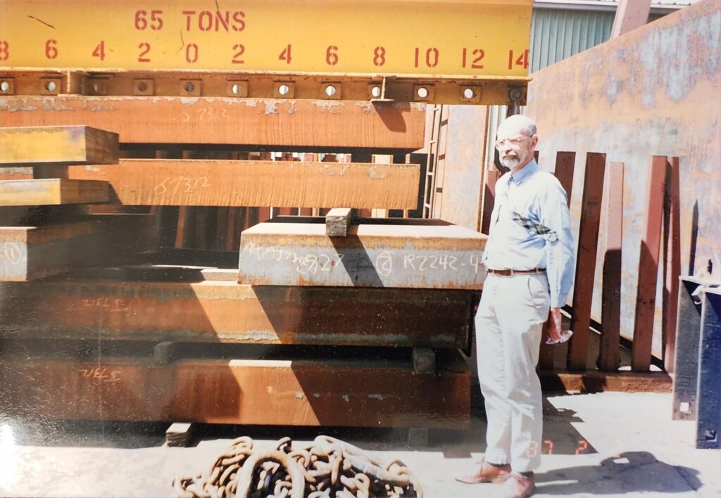 Standing next to a massive stack of large iron slabs and struts, a man gazes off into the distance and out of frame, his tie flapping in what looks to be a strong gust of wind.