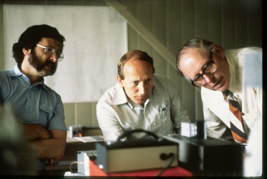 Three men hunch over an apparatus, peering intently at a read-out or monitor that isn't visible from the camera perspective.