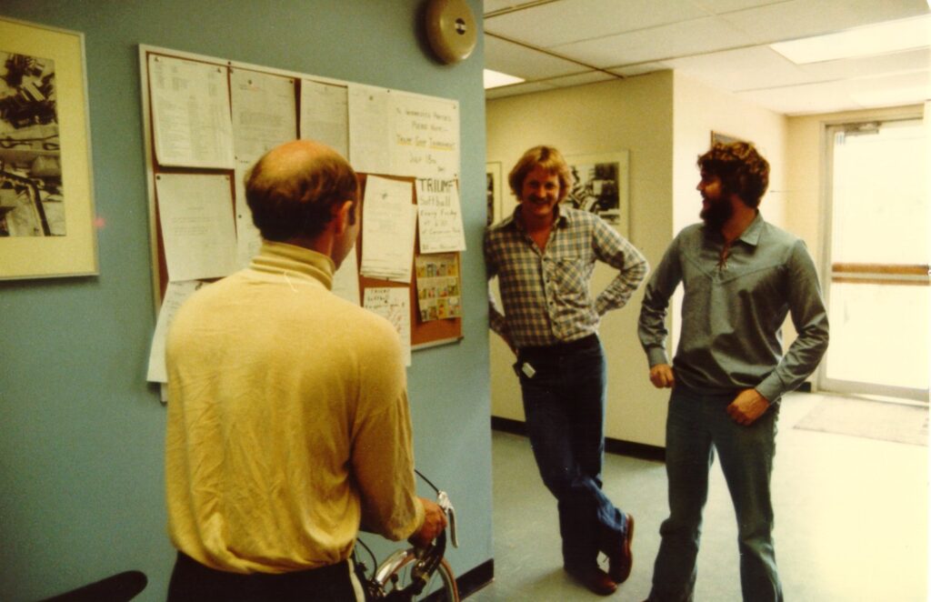 Three men stand at ease in a small group, discussing a set of posted read-outs hanging from a wall nearby.