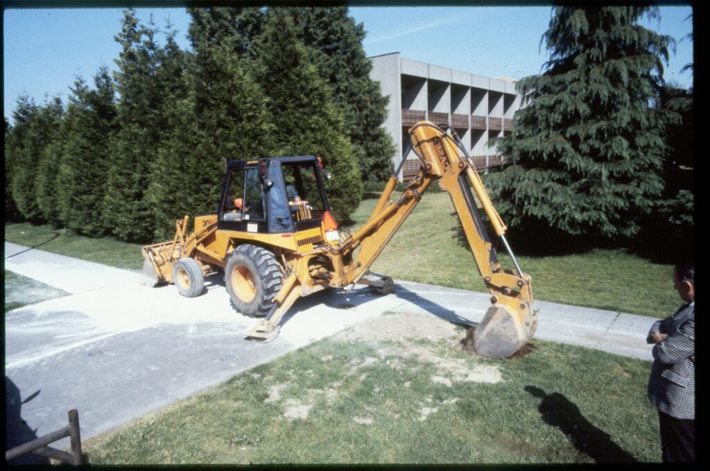 An excavator sits on a sidewalk, its bucket resting next to a small hole.