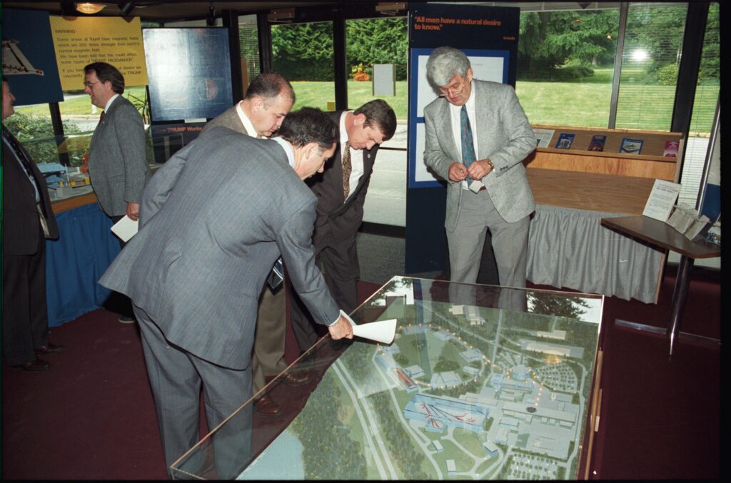 Standing in a dimly-lit lobby, four men bend over to inspect a scale model of the TRIUMF KAON facility.