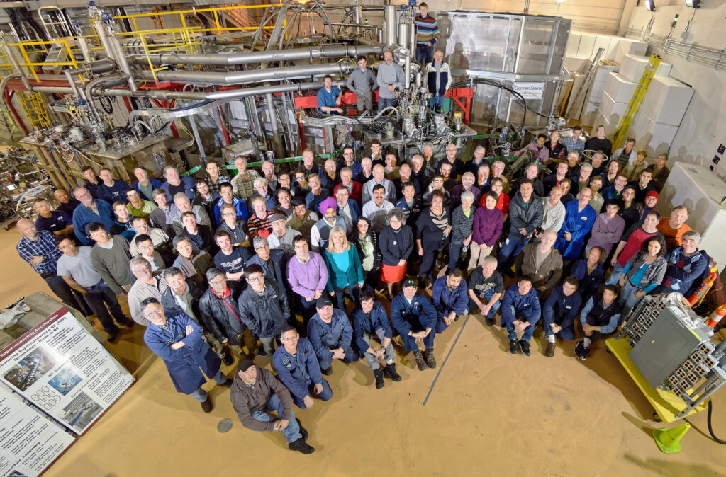 A large group photo of approximately 100 people, with a series of tubes and pipes in the background.