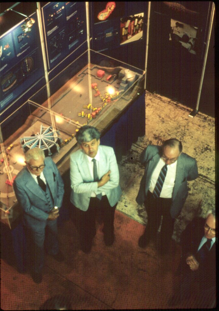 Three men in suits gaze upwards towards the camera, surrounded by public outreach materials and a scale model of the cyclotron facility